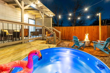 Cozy backyard deck at night with an illuminated hot tub featuring an inflatable flamingo, string lights overhead, and a fire pit surrounded by blue Adirondack chairs.
