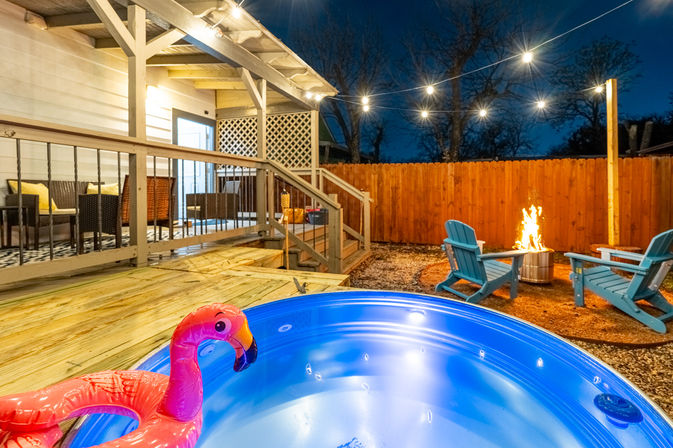 Cozy backyard deck at night with an illuminated hot tub featuring an inflatable flamingo, string lights overhead, and a fire pit surrounded by blue Adirondack chairs.