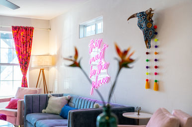 Sunlit boho living room with a teal velvet sofa, pink accent chairs, bright floral curtains, a tripod floor lamp, a neon pink wall sign reading 'this must be the place', a decorative painted bull skull with colorful pom‑pom garland, and blurred tropical flowers in the foreground.