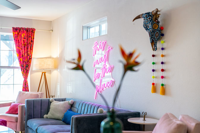 Sunlit boho living room with a teal velvet sofa, pink accent chairs, bright floral curtains, a tripod floor lamp, a neon pink wall sign reading 'this must be the place', a decorative painted bull skull with colorful pom‑pom garland, and blurred tropical flowers in the foreground.