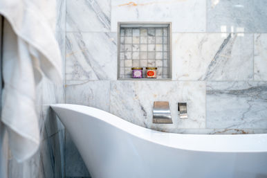 Sleek marble bathroom with a white freestanding tub, chrome wall faucet, recessed tiled niche holding two colorful bath jars and a hanging white towel