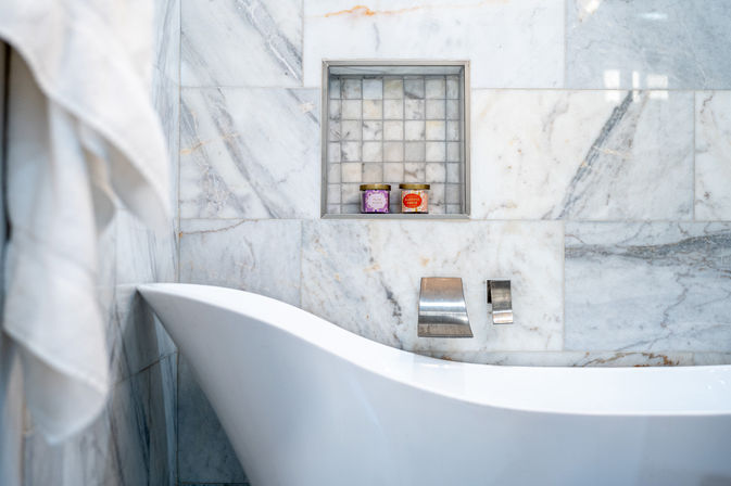 Sleek marble bathroom with a white freestanding tub, chrome wall faucet, recessed tiled niche holding two colorful bath jars and a hanging white towel