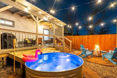 Cozy suburban backyard patio at night: string lights over a galvanized stock‑tank pool with a pink inflatable flamingo, wooden deck seating, Adirondack chairs around a glowing fire pit and privacy fence.