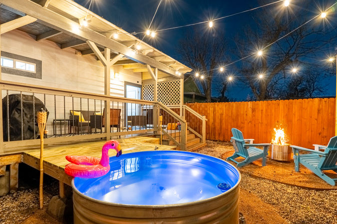 Cozy suburban backyard patio at night: string lights over a galvanized stock‑tank pool with a pink inflatable flamingo, wooden deck seating, Adirondack chairs around a glowing fire pit and privacy fence.