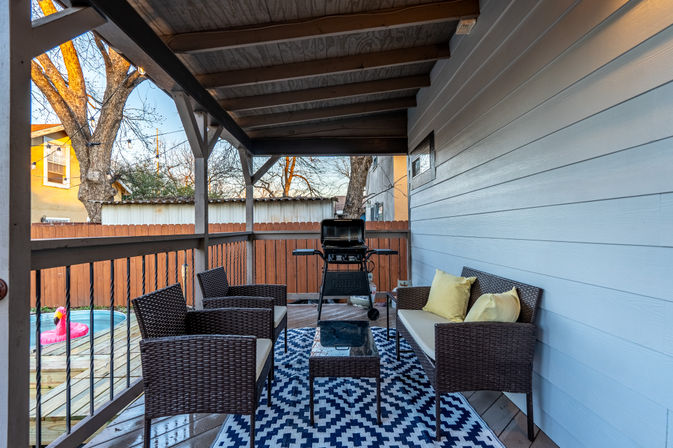 Cozy covered backyard deck with wicker sofa and chairs on a navy geometric rug, gas grill, wooden privacy fence and inflatable pool float in the yard.