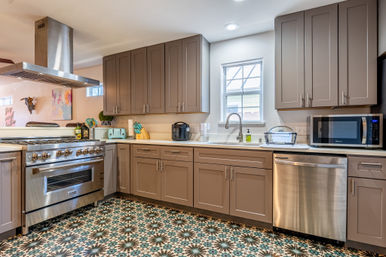 Bright modern kitchen with gray shaker cabinets, stainless steel range, vent hood, dishwasher and microwave, farmhouse sink under a window, mint toaster and bold geometric encaustic-style tile floor.
