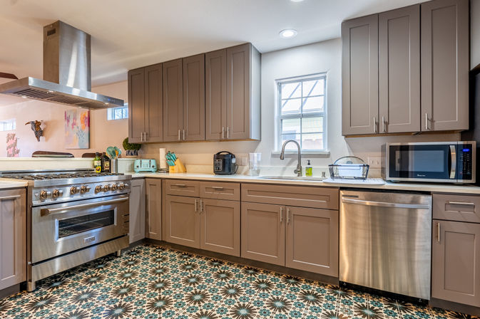 Bright modern kitchen with gray shaker cabinets, stainless steel range, vent hood, dishwasher and microwave, farmhouse sink under a window, mint toaster and bold geometric encaustic-style tile floor.