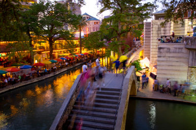 Dusk view of the San Antonio River Walk, Texas — illuminated riverside restaurants with colorful umbrellas, leafy trees and a stone pedestrian bridge with blurred people crossing.