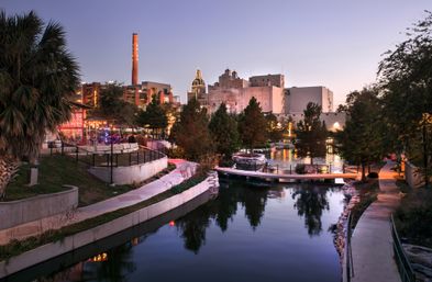 Dusk downtown riverwalk along a calm canal with lit riverside café and pedestrian bridges, palm trees and reflections, and industrial smokestack and historic buildings in the background.