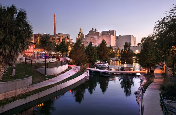 Dusk downtown riverwalk along a calm canal with lit riverside café and pedestrian bridges, palm trees and reflections, and industrial smokestack and historic buildings in the background.