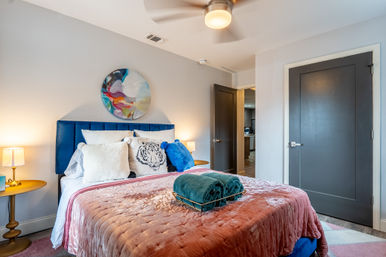 Cozy modern apartment bedroom with blue velvet headboard, pink quilted bedspread, decorative tiger pillow, round abstract wall art, brass bedside lamps, folded green towels, ceiling fan and gray doors.