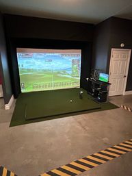 Indoor golf simulator in a garage-style room — large projection screen showing a virtual fairway, green hitting mat with tee and launch monitor, nearby golf club rack and control monitor, concrete floor with black-and-yellow safety tape, ready to tee off
