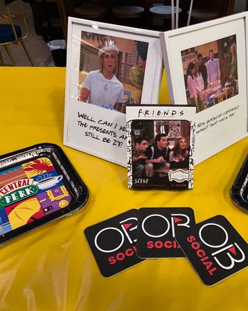 Bright yellow tablecloth display of TV sitcom memorabilia — framed scene photos and a small 'Friends' scene card, a colorful coffee-shop style tray, and three black coasters with a white-and-red logo.