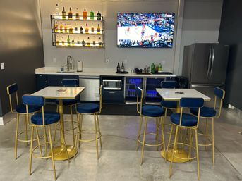 Modern lounge bar area with blue velvet and gold-frame bar stools around marble-top high tables, backlit liquor shelves, wall-mounted TV showing a basketball game, compact kitchenette and stainless refrigerator.