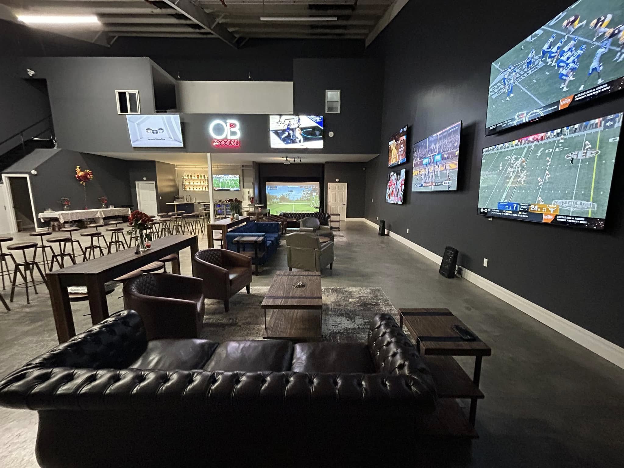 Spacious industrial sports lounge with tufted leather sofas, armchairs, high-top tables and a bar area facing multiple large wall TVs and a projection screen showing football — perfect for watch parties.