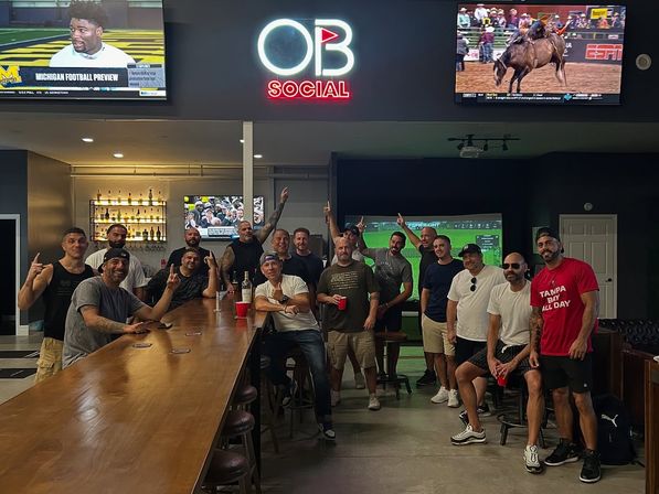 Group of men posing in a modern sports bar interior with a long wooden bar, neon sign, stools and multiple TVs showing football and rodeo highlights.
