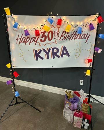 Festive indoor 30th birthday banner reading 'KYRA' with twinkling string lights, colorful plastic-cup garland and stacked gift bags on the floor.