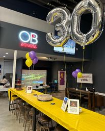 Indoor event space set up for a 30th birthday — large silver 30 balloons, yellow and purple balloons, long table with yellow tablecloth, cake and bar seating.