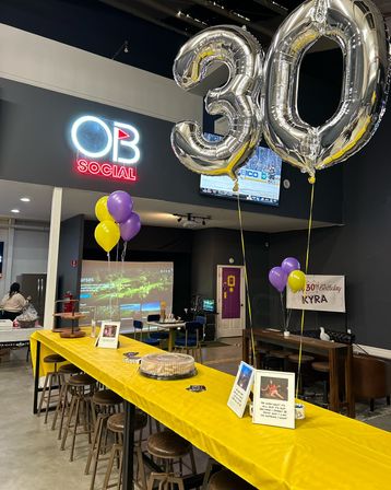 Indoor event space set up for a 30th birthday — large silver 30 balloons, yellow and purple balloons, long table with yellow tablecloth, cake and bar seating.