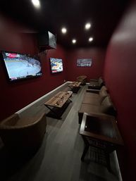 Cozy long home media room with burgundy walls, recessed lights, two wall-mounted TVs showing sports, brown leather sofa and chairs, wooden coffee tables and gray wood floor.
