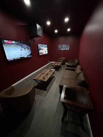 Cozy long home media room with burgundy walls, recessed lights, two wall-mounted TVs showing sports, brown leather sofa and chairs, wooden coffee tables and gray wood floor.