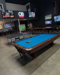 Indoor sports bar and game lounge with a blue-felt pool table racked and ready, adjacent ping-pong table, multiple wall-mounted TVs showing sports, a neon sign and long bar with stools on a concrete floor.