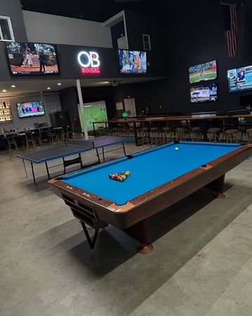 Indoor sports bar and game lounge with a blue-felt pool table racked and ready, adjacent ping-pong table, multiple wall-mounted TVs showing sports, a neon sign and long bar with stools on a concrete floor.