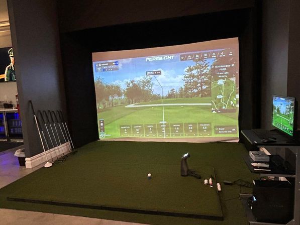 Indoor golf simulator bay with green hitting mat, clubs leaning against a wall, projector screen showing a virtual fairway and side monitor, golf balls and tee ready for a virtual tee shot.