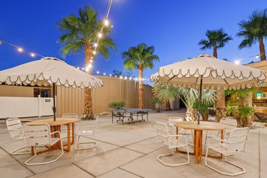 Outdoor patio at dusk with scalloped umbrellas, round wooden tables, white metal chairs, string lights, palm trees and a ping-pong table