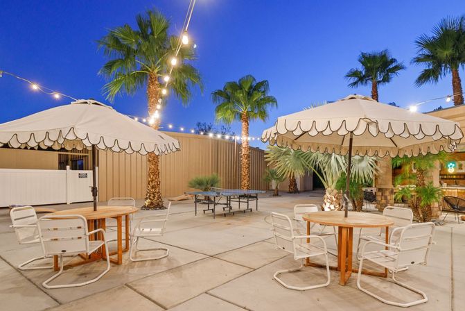 Outdoor patio at dusk with scalloped umbrellas, round wooden tables, white metal chairs, string lights, palm trees and a ping-pong table