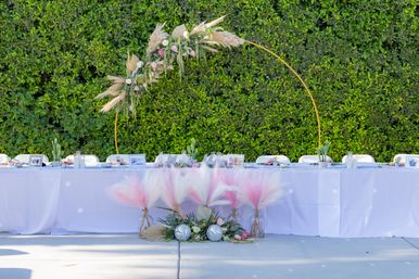 Outdoor garden wedding head table with white linens and a gold circular arch adorned with pampas grass and blush roses, pink-and-white pampas plumes in vases, disco ball accents and a lush green hedge backdrop.