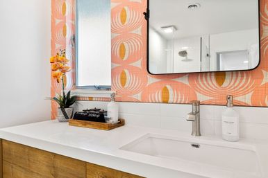 Bright modern bathroom vanity with white quartz countertop and undermount sink, wood cabinets, chrome faucet, coral retro-pattern wallpaper, black-framed mirror, frosted window, potted yellow orchid and wooden tray with soap and makeup pouch
