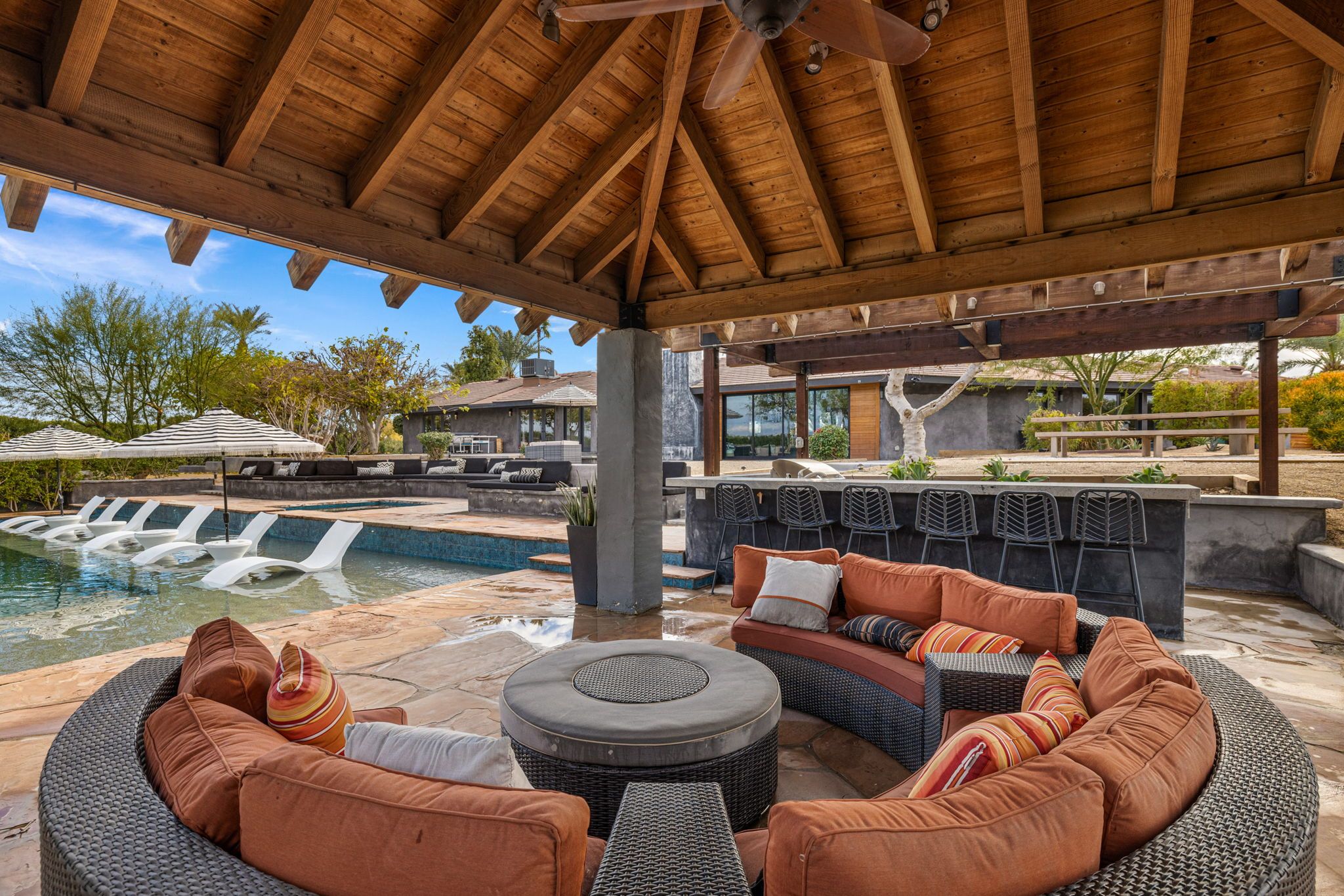 Covered wooden pavilion with curved wicker sofa and orange cushions around a round table, adjacent pool with in-pool loungers and striped umbrellas, outdoor bar seating and modern home — luxury backyard poolside lounge.