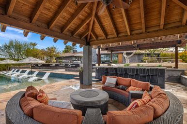 Covered wooden pavilion with curved wicker sofa and orange cushions around a round table, adjacent pool with in-pool loungers and striped umbrellas, outdoor bar seating and modern home — luxury backyard poolside lounge.