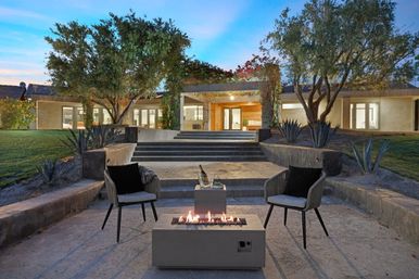 Modern suburban backyard patio at dusk with concrete fire pit, two lounge chairs, champagne on a side table, agave plants and steps leading to an illuminated contemporary home