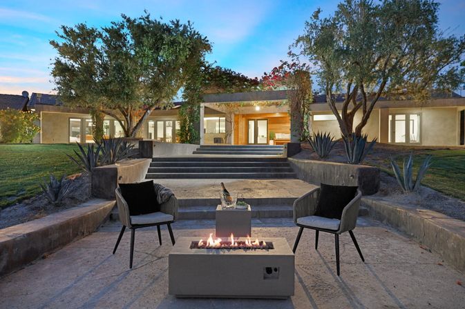 Modern suburban backyard patio at dusk with concrete fire pit, two lounge chairs, champagne on a side table, agave plants and steps leading to an illuminated contemporary home