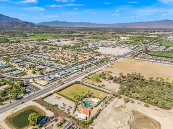 Aerial view of a desert suburban community with rows of single-story homes, intersecting roads, palm tree orchards and open farmland stretching toward distant mountains under a bright blue sky.