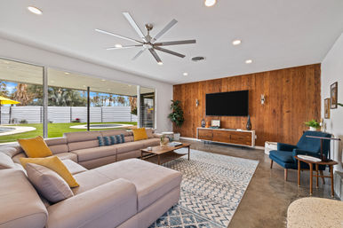 Bright mid-century modern living room with large beige sectional and yellow pillows, wood-paneled TV wall, oversized ceiling fan, patterned area rug, and sliding glass wall opening to a grassy backyard with circular pool and palm trees.