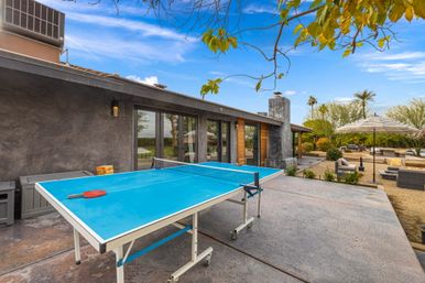 Bright blue outdoor ping-pong table on a modern desert-style backyard patio with lounge seating, umbrella, palm trees and a clear blue sky