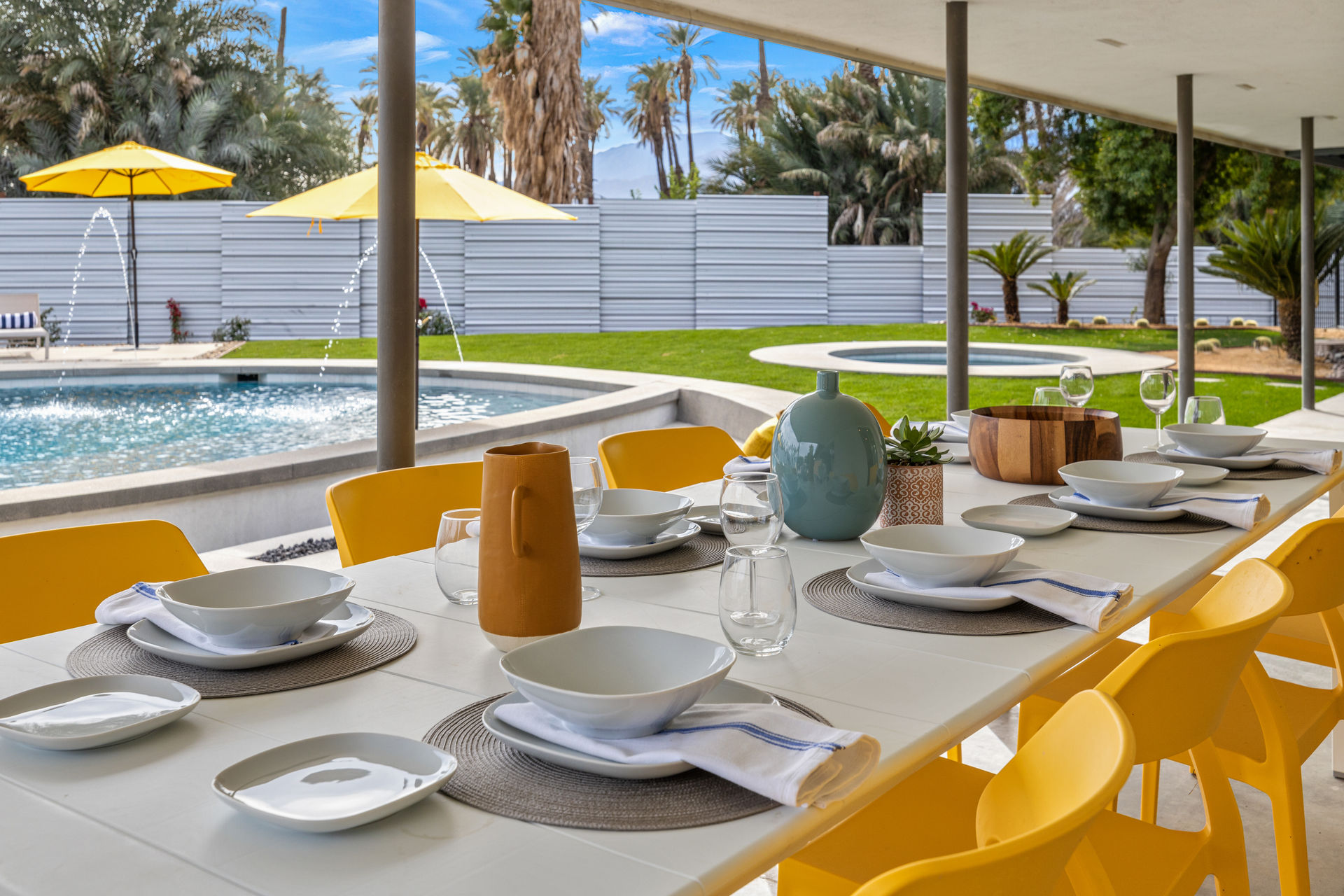 Sunny outdoor dining table with white dishes and yellow chairs on a covered patio overlooking a resort-style pool, yellow umbrellas and palm trees in a mid-century modern backyard.
