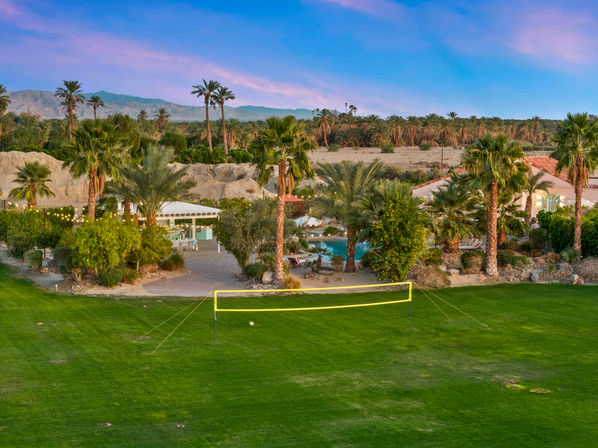 Dusk desert oasis with a yellow volleyball net and ball on a green lawn, palm trees surrounding a lit pool and patio, rocky hills and mountains under a pink-blue sky.