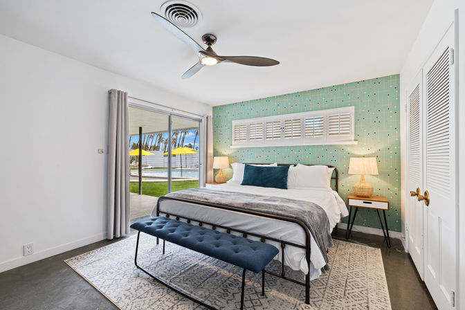 Sunny coastal-style bedroom with king bed, teal mid-century patterned accent wall, ceiling fan, navy tufted bench and bedside lamps; sliding glass door opens to lawn, pool and yellow umbrellas.