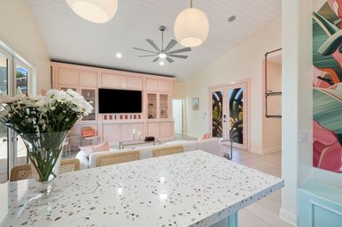 Bright sunlit open-plan living room and kitchen with terrazzo island in the foreground, vase of white daisies, pastel pink built-in media wall with TV, glass French doors, modern ceiling fan and pendant lights.
