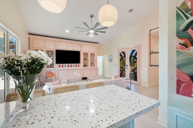 Bright sunlit open-plan living room and kitchen with terrazzo island in the foreground, vase of white daisies, pastel pink built-in media wall with TV, glass French doors, modern ceiling fan and pendant lights.