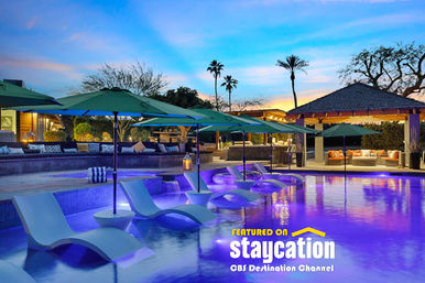 Twilight resort pool with in-water lounge chairs and green umbrellas, purple LED-lit water, palm trees and a cozy cabana seating area.