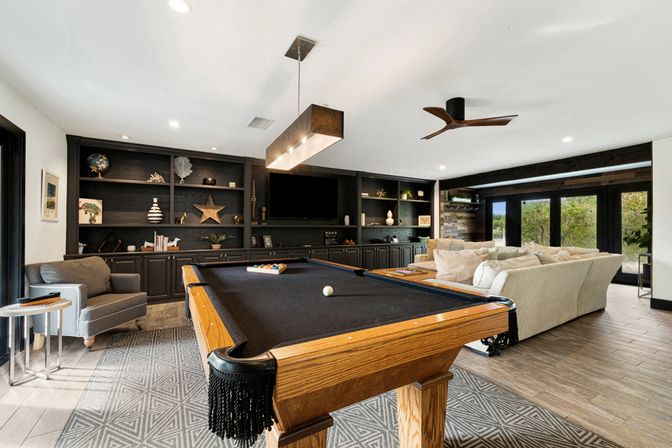 Contemporary game room with wooden pool table (black felt), cozy beige sectional, dark built-in shelving and TV, pendant light, ceiling fan, and large glass doors with yard views.