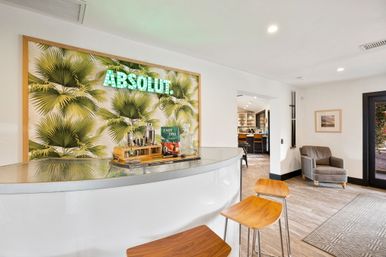 Modern home bar with curved white counter and wooden stools, tropical palm wallpaper backlit by a green neon sign, cocktail tools and a tiki cocktail book on the bar, open kitchen and cozy gray armchair in a bright living area