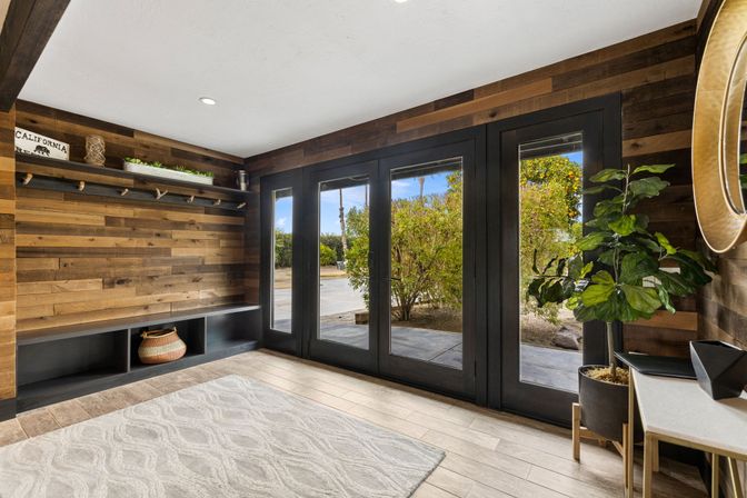 Modern rustic California entryway mudroom with reclaimed wood accent wall, black-framed glass French doors to the yard, built-in bench storage with woven basket, potted fiddle-leaf fig, and light patterned rug.