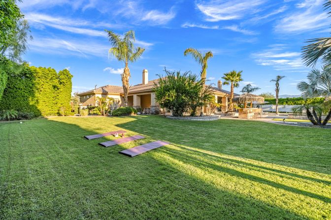 Sunny suburban backyard with manicured green lawn, three purple yoga mats, tall palm trees, single-story stucco house with tiled roof, patio seating and a poolside tiki bar