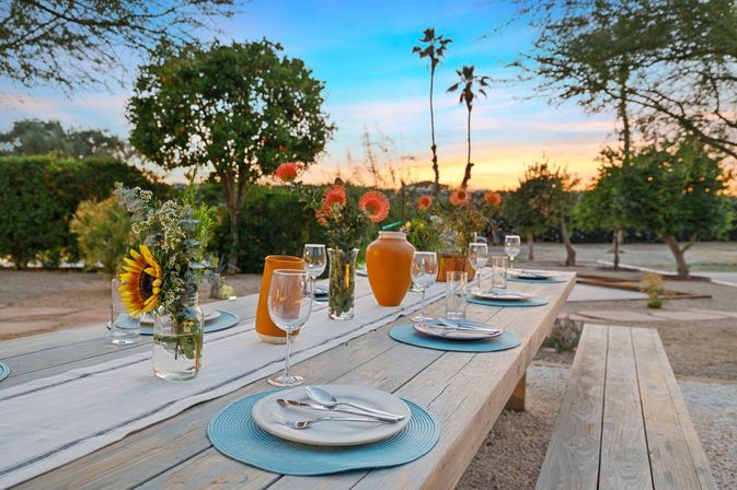 Outdoor wooden dining table set for a sunset dinner in a garden — blue placemats, white plates and cutlery, wine glasses, sunflower in a mason jar and orange vases, benches with palm trees and a colorful sky in the background.